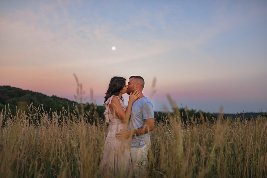 Couple standing in a field of tall grass gazing at one another and kissing under a cotton candy sky and a Full Moon