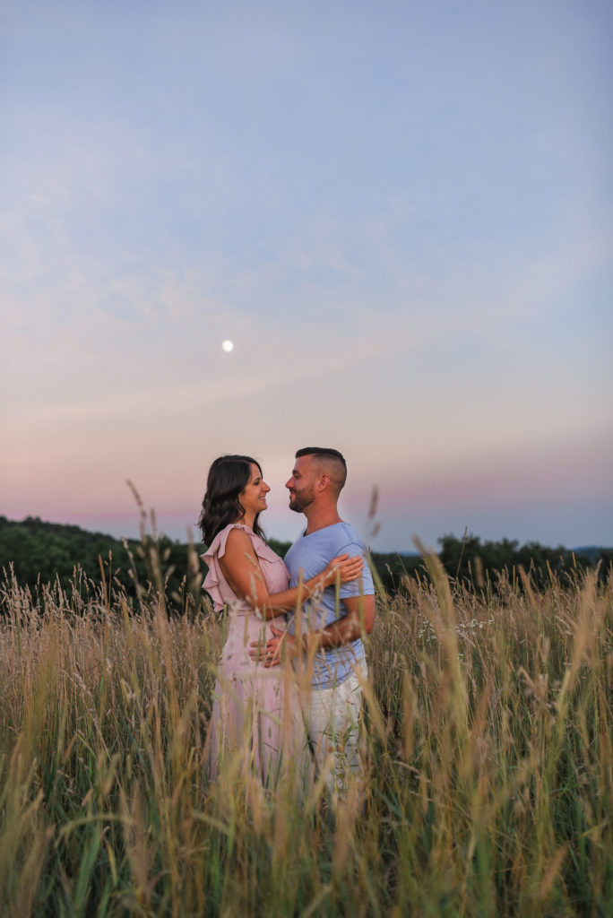 Location of engagement session matters.Couple standing in a field of tall grass gazing at one another and kissing under a cotton candy sky and a Full Moon
