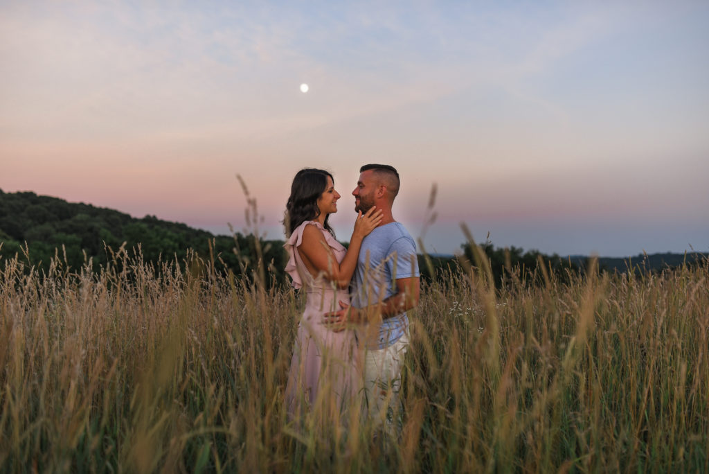 Location of engagement session matters.Couple standing in a field of tall grass gazing at one another and kissing under a cotton candy sky and a Full Moon